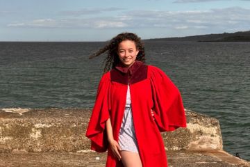 A young, mixed race, female student, wearing a red University of St Andrews undergraduate gown, smiling and standing on the end of a pier with the sea in the background